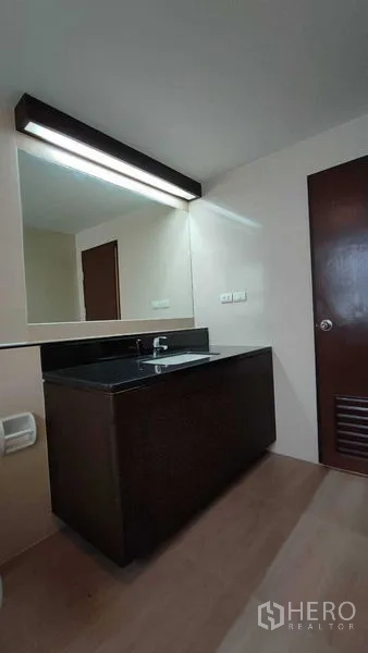 Condominium for rent in Khlong Toei, Bangkok - Vanity area with long backlit mirror and dark countertop in the condo bathroom.