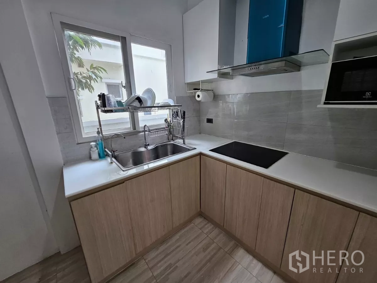 Detached House for rent in Khlong Toei, Bangkok - Modern L-shaped kitchen with window over sink, induction hob, hood and light wood cabinets.