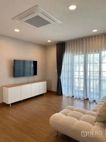 Detached House for rent in Bang Phli, Samut Prakan - Bright living room with wall-mounted TV, white console, sheer curtains and wood flooring.