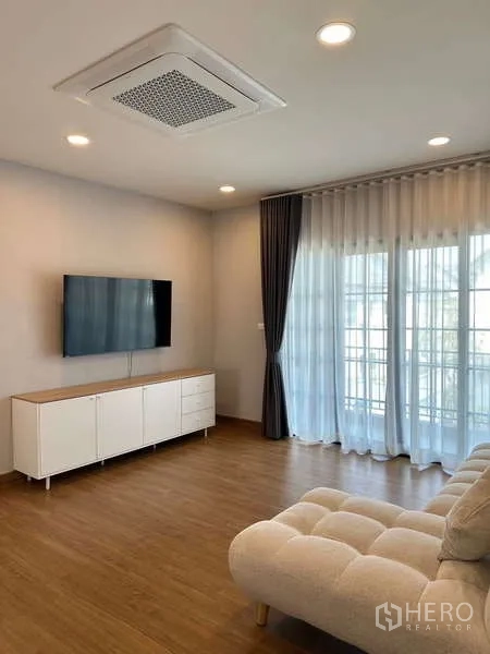 Detached House for rent in Bang Phli, Samut Prakan - Bright living room with wall-mounted TV, white console, sheer curtains and wood flooring.