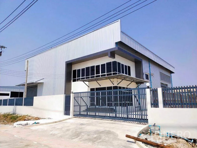 Factory for rent in Samut Prakan - Modern metal-clad factory exterior with corner office windows, sliding steel gate, concrete driveway, and a high roller shutter door.