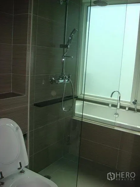 Condominium for rent in Khlong Toei, Bangkok - Modern bathroom with glass shower, rainfall head and bathtub beside a frosted window.