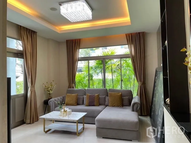 Detached House for sale in Lat Krabang, Bangkok - Bright family lounge with L-shaped sofa, floor-to-ceiling windows and tray ceiling lights.