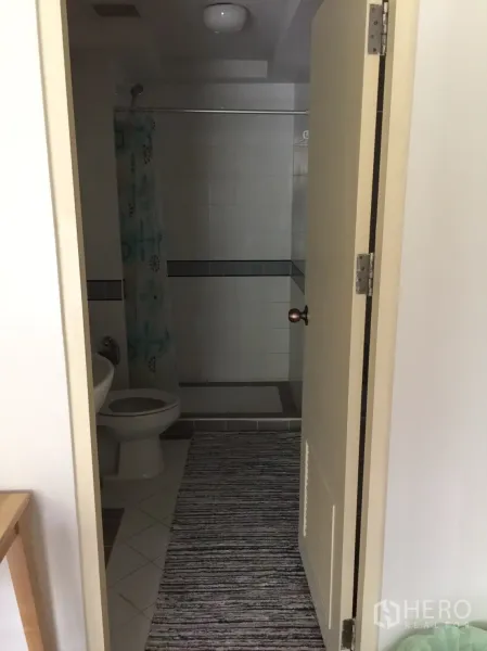 Condominium for rent in Khlong Toei, Bangkok - Bathroom viewed from the doorway showing toilet, sink, and tiled shower with curtain.