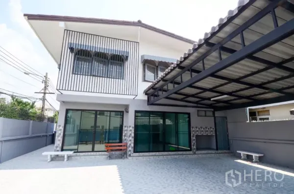 带办公室的家 出租在Lat Phrao, Bangkok - 两层转角住办合一建筑，带遮棚车位、推拉玻璃门与铺砖前院。