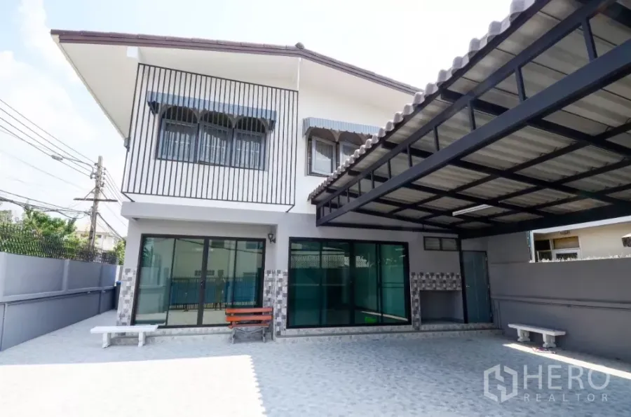 带办公室的家 出租在Lat Phrao, Bangkok - 两层转角住办合一建筑，带遮棚车位、推拉玻璃门与铺砖前院。