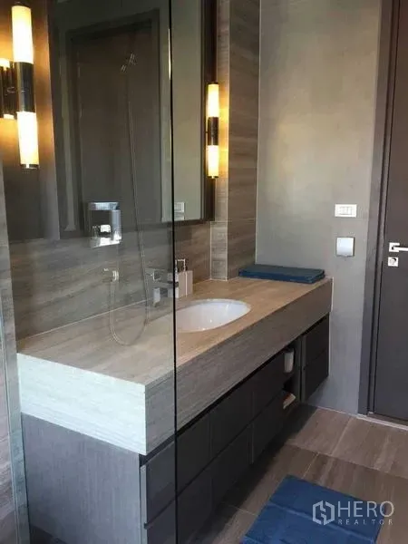 Condominium for rent in Bang Rak, Bangkok - Contemporary bathroom with long vanity, glass shower enclosure, and warm lighting.