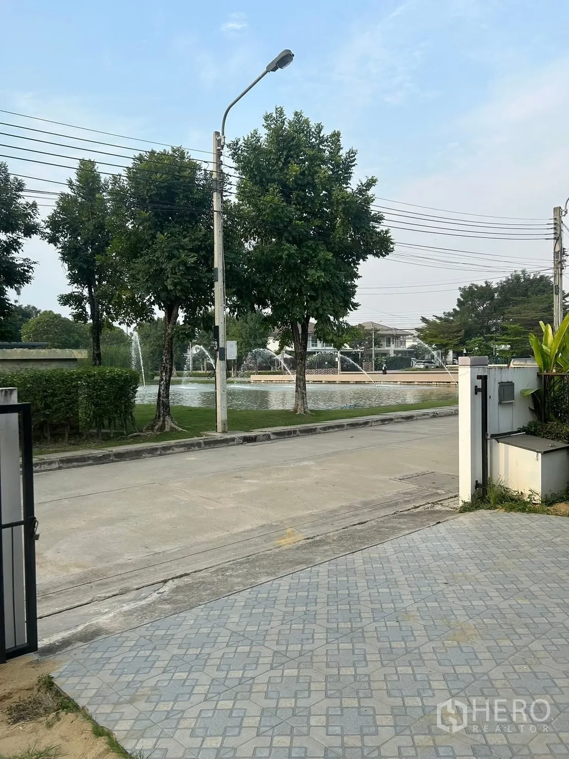 Detached House for rent in Bang Na, Bangkok - Front gate and tiled driveway facing a lakeside street with trees and fountains.