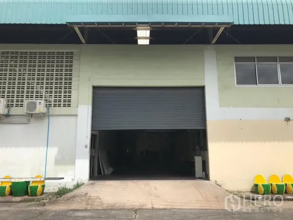 货栈 出租在Bang Phli, Samut Prakan - 近景卷帘货门与混凝土坡道，雨棚宽阔并设有窗户。