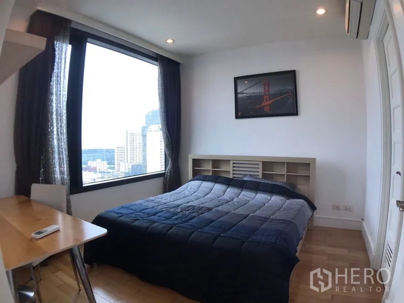 Condominium for rent in Khlong Toei, Bangkok - Bedroom with large window and city view, desk, air-conditioning and wooden floor with a dark duvet on the bed.