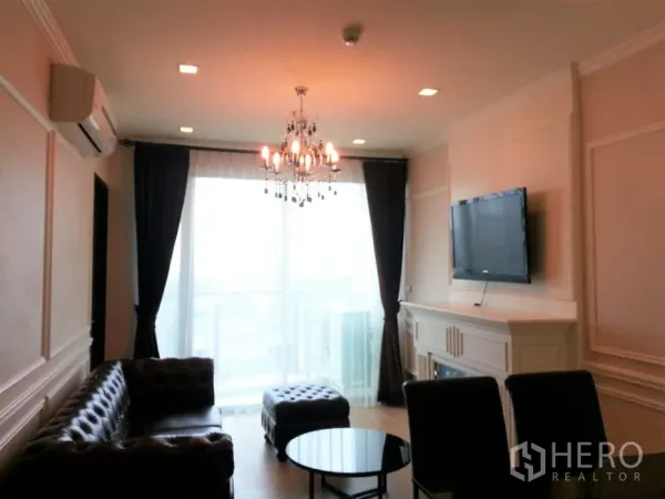 Condominium for rent or sale in Watthana, Bangkok - Living area view of chandelier, dark curtains, wall-mounted TV and round coffee table by balcony glazing.