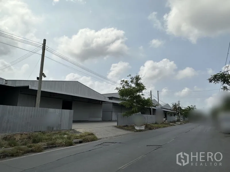 Warehouse for rent in Mueang Samut Prakan, Samut Prakan - Street view of a row of steel warehouses along a quiet road with utility lines and trees.