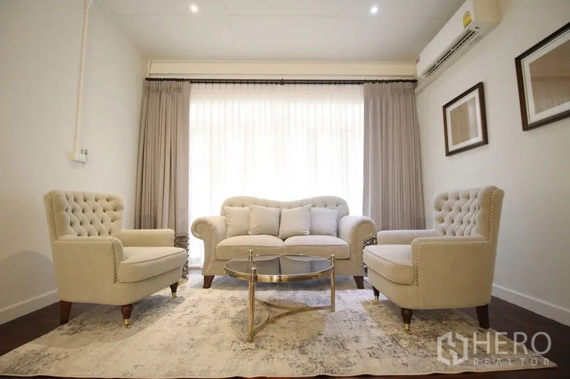 Detached House for rent in Chatuchak, Bangkok - Cozy sitting area with tufted beige sofa and armchairs, glass coffee table, rug, sheer curtains, and air conditioner.