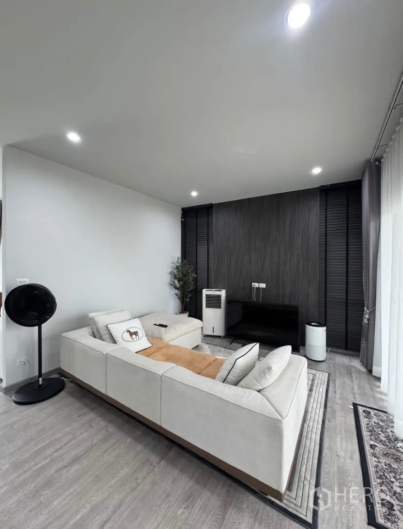 Detached House for rent or sale in Lat Krabang, Bangkok - Contemporary living room with L-shaped sofa, recessed lighting, dark feature wall, and media console.