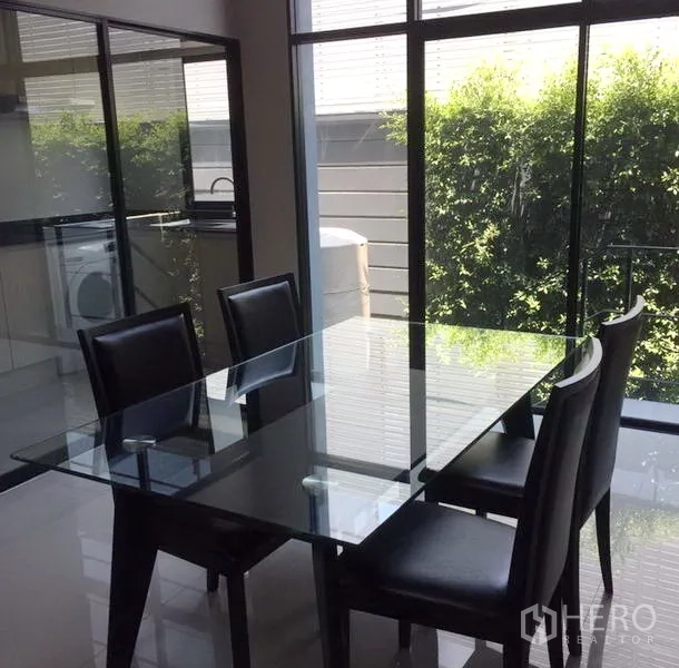 Townhouse for rent in Suan Luang, Bangkok - Bright dining area with a glass table and black chairs beside floor-to-ceiling windows overlooking greenery.