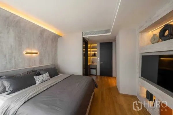 Condominium for sale in Khlong Toei, Bangkok - Primary bedroom with curved concrete-style feature wall, warm cove lighting, king bed, and built-in TV shelving.