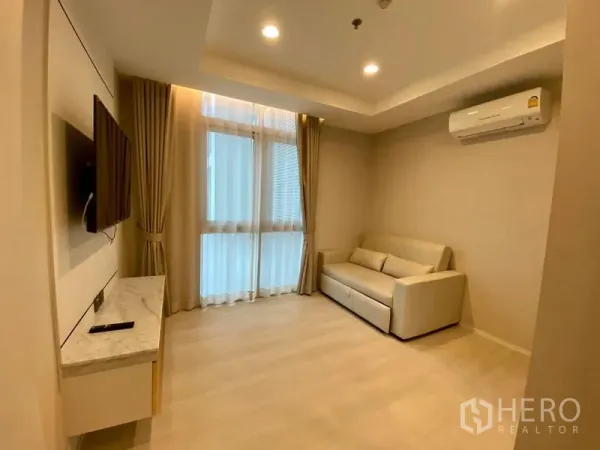 Condominium for rent in Khlong Luang, Pathum Thani - Cozy multi-purpose room with sofa, wall-mounted TV, tall curtained windows and air conditioning.