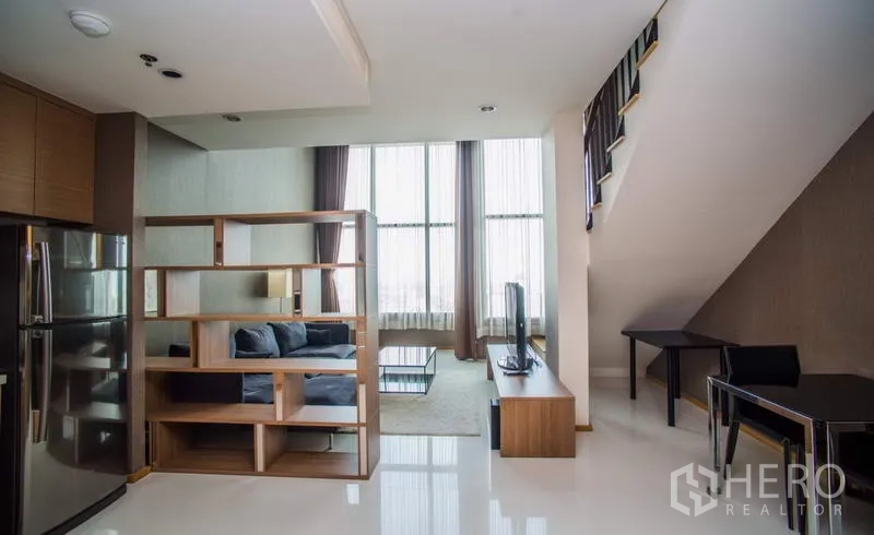 Condominium for rent or sale in Khlong Toei, Bangkok - Living area with floor-to-ceiling windows, shelving divider, and stairs to mezzanine.