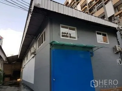 คลังสินค้า ให้เช่าในChatuchak, Bangkok - ภายนอกโกดังสีเทาในเขตจตุจักร ประตูบานเลื่อนเหล็กสีน้ำเงิน หน้าต่างเล็ก มีเครื่องปรับอากาศ และหลังคาเมทัลชีท ใกล้อาคารสูง