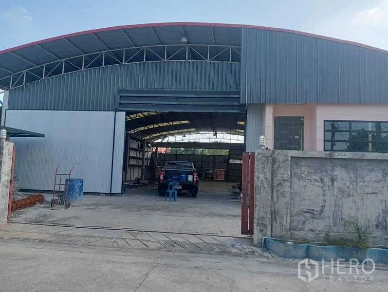 Warehouse for rent in Lam Luk Ka, Pathum Thani - Gated entrance to warehouse showing open loading bay, pickup truck and spacious concrete yard.