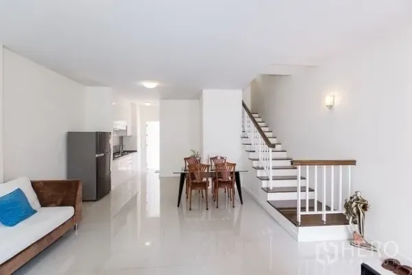 Townhouse for rent in Watthana, Bangkok - Spacious open-plan layout showing dining set, sofa and wood-trim staircase leading to upper floors.