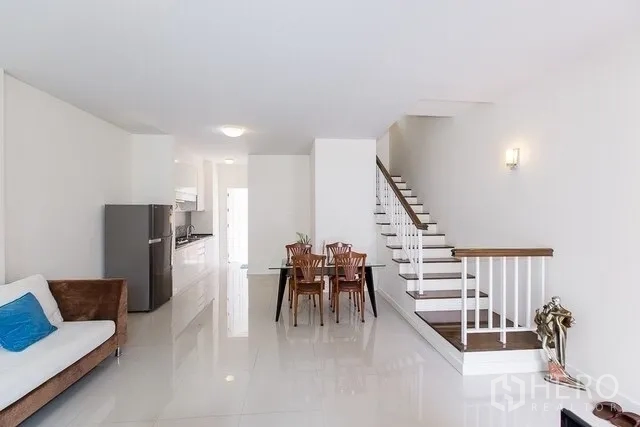 Townhouse for rent in Watthana, Bangkok - Spacious open-plan layout showing dining set, sofa and wood-trim staircase leading to upper floors.