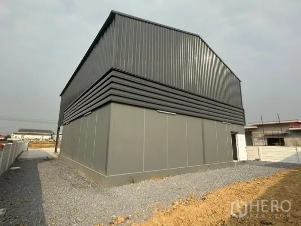 Factory for rent or sale in Bang Bua Thong, Nonthaburi - Rear and side of steel‑clad warehouse with ventilation bands and gravel service path.