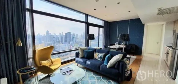 Condominium for sale in Ratchathewi, Bangkok - Open-plan lounge with navy sofa, large corner windows, and city view beside compact kitchen and dining.