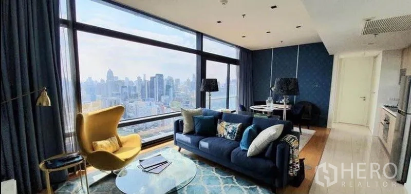 Condominium for sale in Ratchathewi, Bangkok - Open-plan lounge with navy sofa, large corner windows, and city view beside compact kitchen and dining.