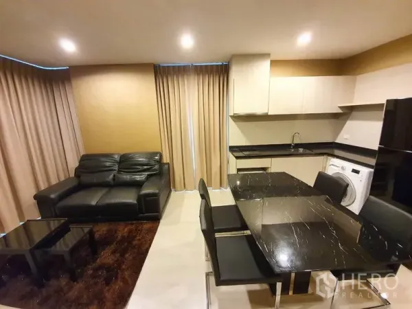 Condominium for rent in Sathon, Bangkok - Open-plan living and kitchen with black stone dining table, leather sofa, beige curtains, and built-in cabinetry with washer and fridge.