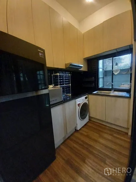 Townhouse for rent in Suan Luang, Bangkok - Fitted kitchen with light wood cabinetry, black countertops, window, fridge and washer.