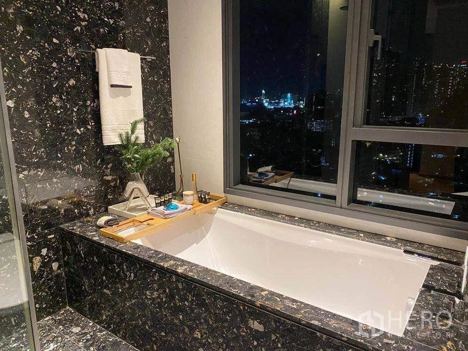 Condominium for rent or sale in Khlong Toei, Bangkok - Soaking bathtub framed by dark stone tiles with a window and night city view.