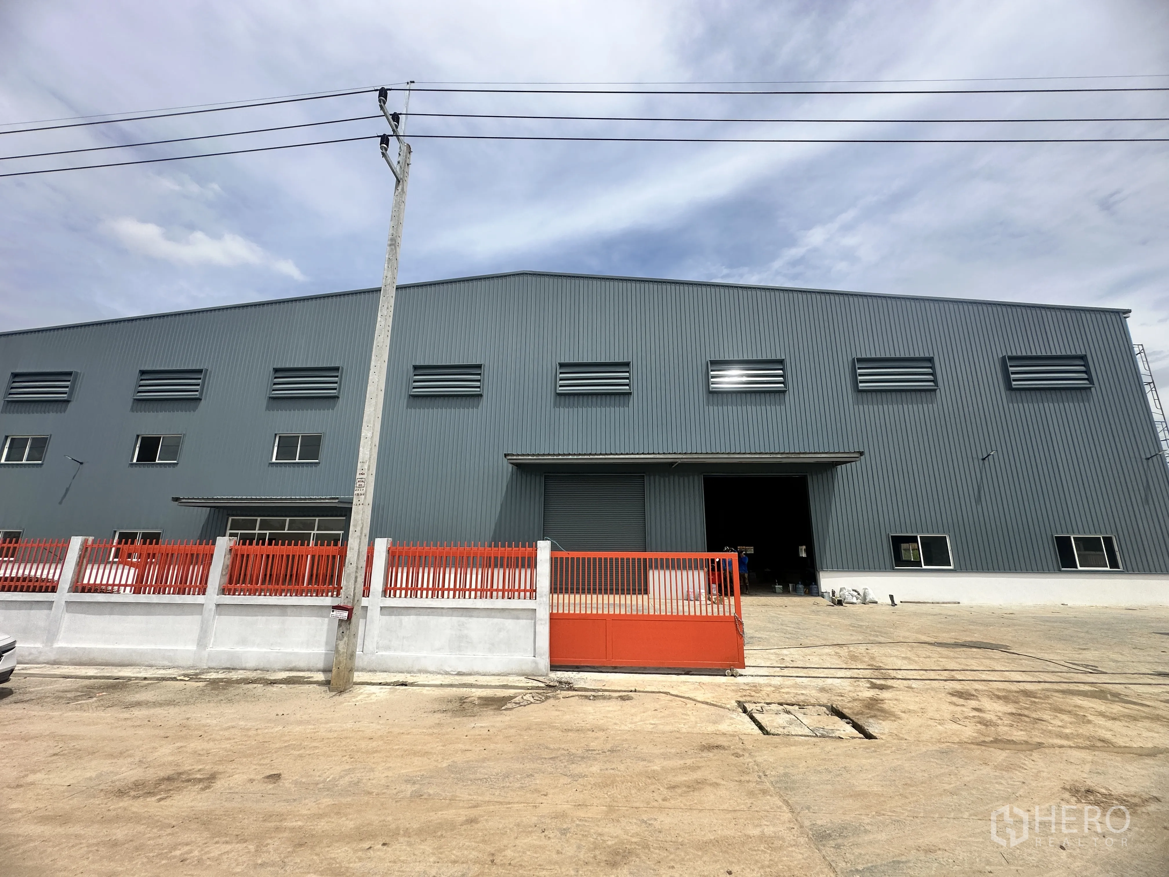 Warehouse for rent or sale in Lam Luk Ka, Pathum Thani - Front elevation of the warehouse behind a sliding gate with a driveway.