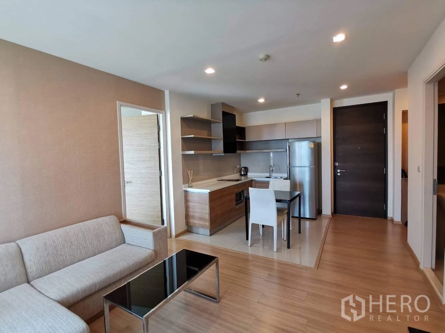 Condominium for rent or sale in Khlong Toei, Bangkok - Open-plan living room with an L-shaped sofa, black glass coffee table, and compact kitchen with shelving and a stainless-steel fridge.