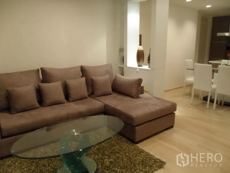 Condominium for rent in Khlong Toei, Bangkok - L-shaped sectional sofa with cushions and oval glass coffee table on a rug in a neutral-toned lounge.