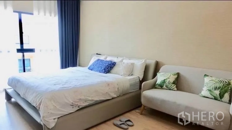 Townhouse for rent or sale in Suan Luang, Bangkok - Spacious bedroom with a bed and small sofa opening to a balcony with curtains.