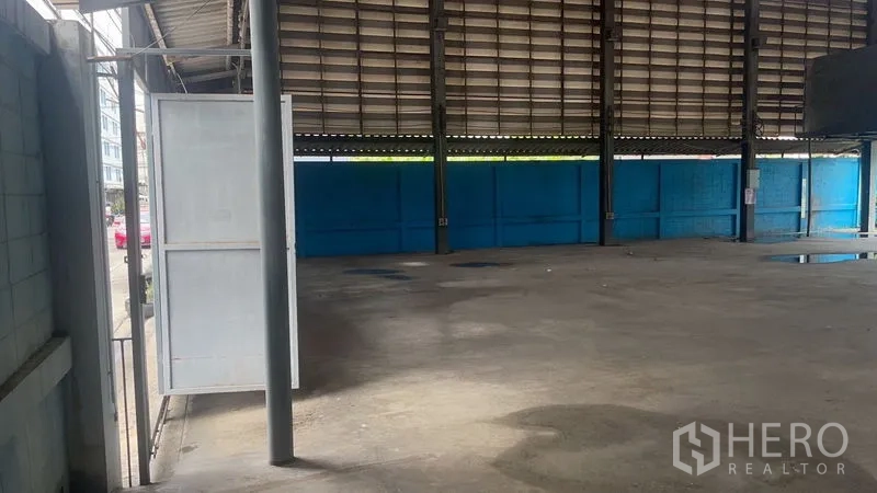 Warehouse for rent in Bang Na, Bangkok - Open-plan warehouse interior with high steel roof, concrete floor, blue perimeter wall and a side swing door.