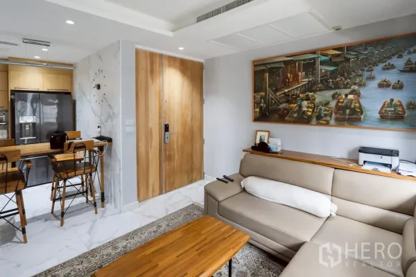 公寓 出租在Pathum Wan, Bangkok - 明亮客厅，米色L形沙发、木质茶几，入户门与餐吧及大理石地面。