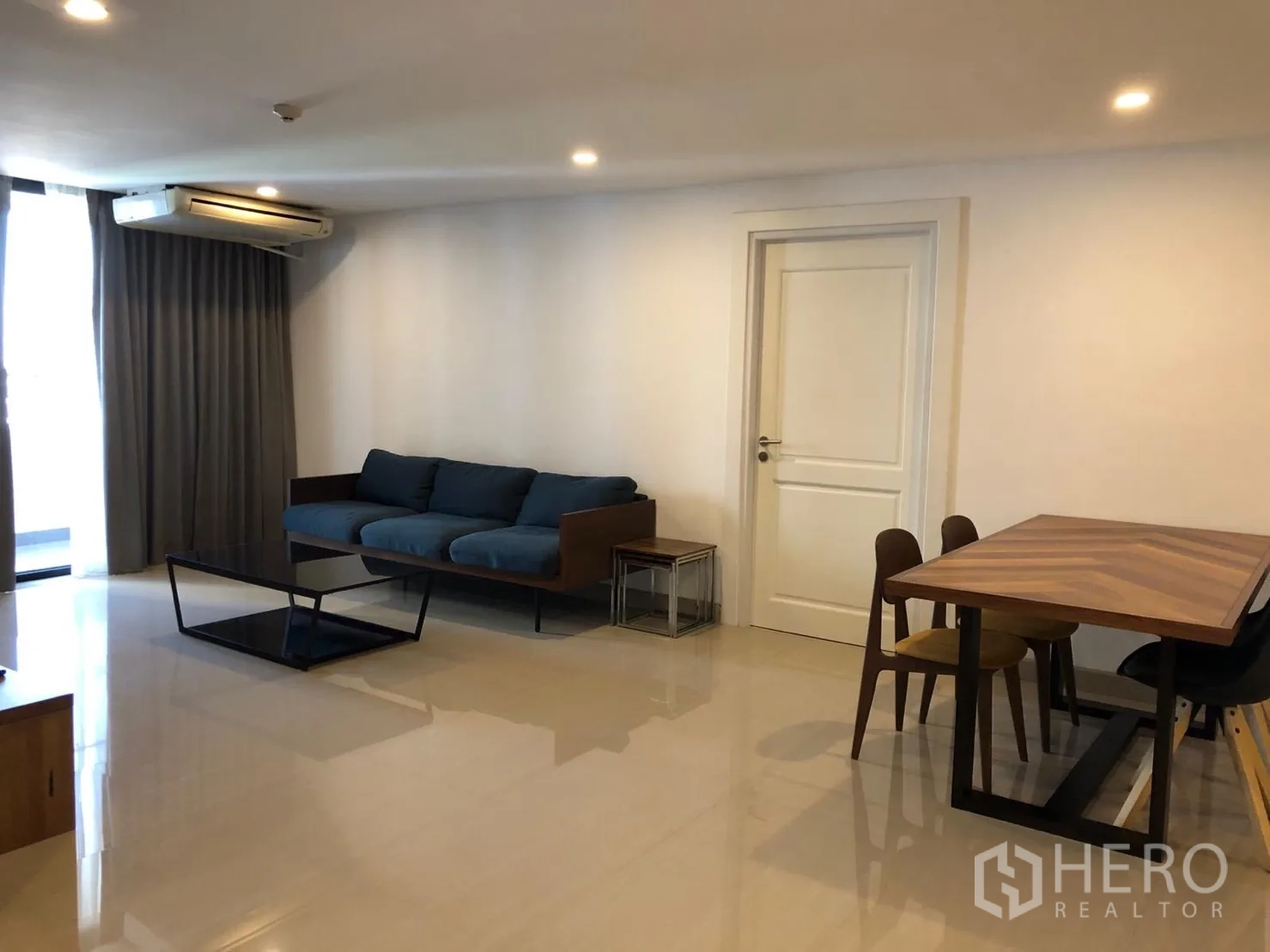 公寓 出租在Watthana, Bangkok - 宽敞客餐厅，蓝色沙发、木质餐桌、空调与落地窗帘