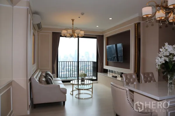 Condominium for rent in Huai Khwang, Bangkok - Open-plan lounge and dining area with city-view balcony, wall-mounted TV, marble-top dining island, upholstered chairs and chandeliers.