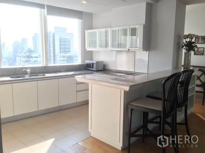 Condominium for rent in Khlong Toei, Bangkok - Sunlit kitchen with white cabinetry, island breakfast bar and large windows facing the city skyline.
