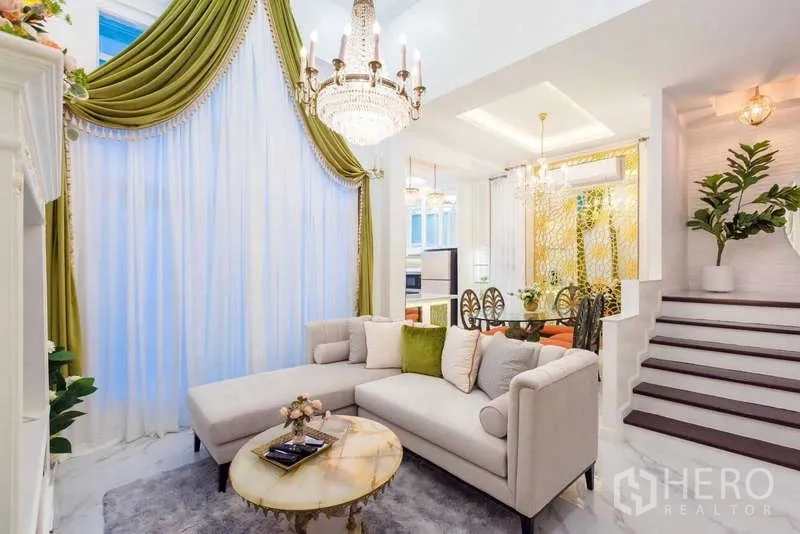 Townhouse for rent in Lat Phrao, Bangkok - Elegant living room with crystal chandelier, tall drapes, L-shaped sofa and open dining area.