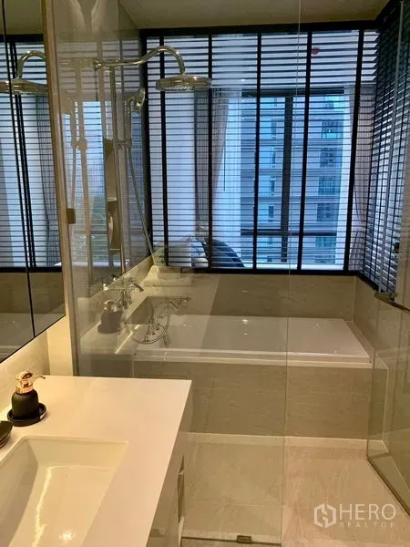 Condominium for rent in Pathum Wan, Bangkok - Modern bathroom with glass shower, bathtub, and large window with blinds.