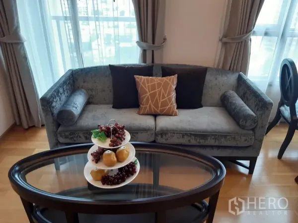 公寓 出租在Watthana, Bangkok - 灰色天鹅绒沙发靠窗，椭圆茶几摆放水果装饰。