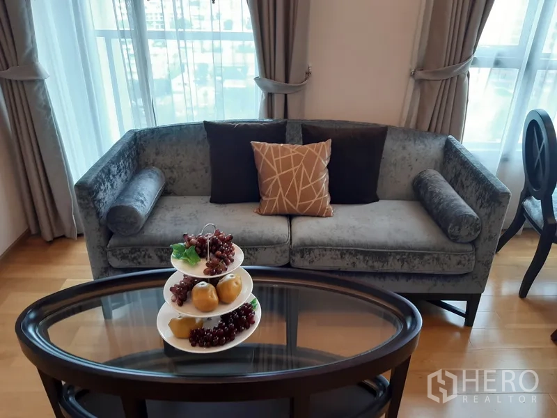 公寓 出租在Watthana, Bangkok - 灰色天鹅绒沙发靠窗，椭圆茶几摆放水果装饰。