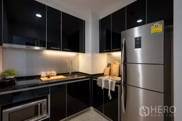 Condominium for rent or sale in Khlong Toei, Bangkok - Modern U-shaped kitchen with black gloss cabinets, under-cabinet lighting and a stainless refrigerator.