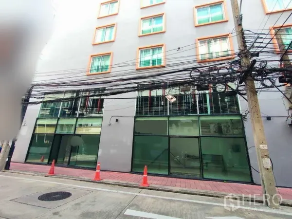Retail Space for rent in Watthana, Bangkok - Ground-floor retail frontage with full-height glass beneath a mid-rise building, red-tile sidewalk, traffic cones, and overhead utility cables.
