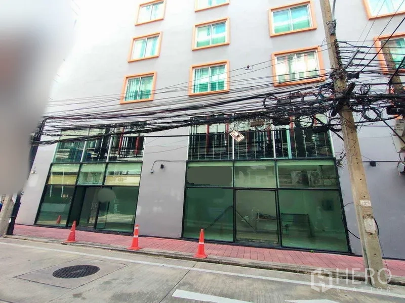 Retail Space for rent in Watthana, Bangkok - Ground-floor retail frontage with full-height glass beneath a mid-rise building, red-tile sidewalk, traffic cones, and overhead utility cables.