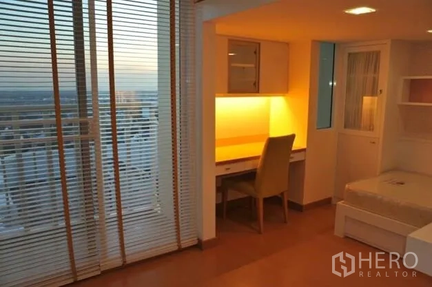公寓 出租或出售在Watthana, Bangkok - 卧室角落设学习桌，暖色灯光、百叶帘和夕阳阳台景。