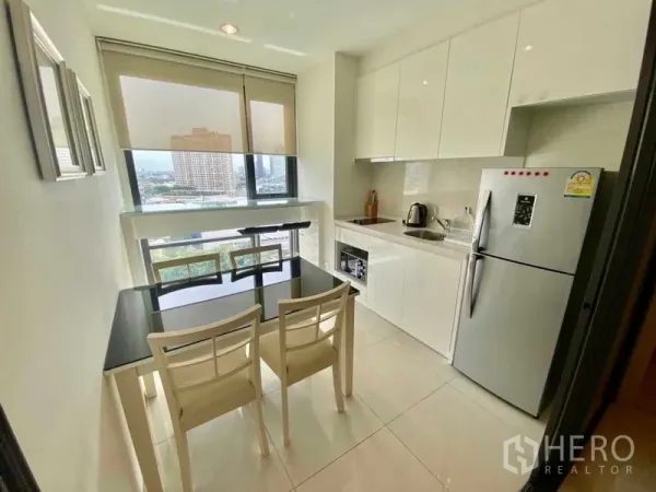 Condominium for rent in Khlong Toei, Bangkok - White kitchen with built-in appliances, a two-seat dining table and a large window with city views.